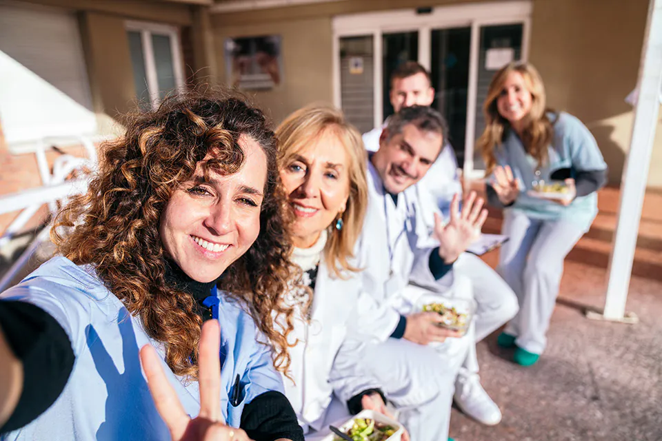 Fünf Personen in medizinischer Kleidung sitzen draußen auf einer Bank und machen ein Selfie, eine Person zeigt ein Peace-Zeichen, eine andere Person hält die Hand wie zum Gruß