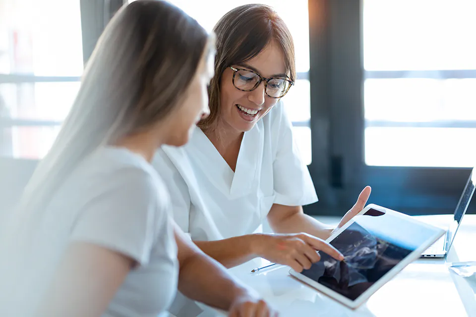 Arbeiten bei RH Zusammenarbeit am Tablet: Eine Fachkraft zeigt der anderen Befunde direkt auf dem Bildschirm, im Hintergrund ein Laptop an einem hellen Arbeitsplatz.