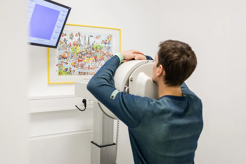 Mann sitzt mit dem Kopf vor einem Gerät für bildgebende Diagnostik, daneben Monitor und ein buntes Wimmelbild an der Wand.