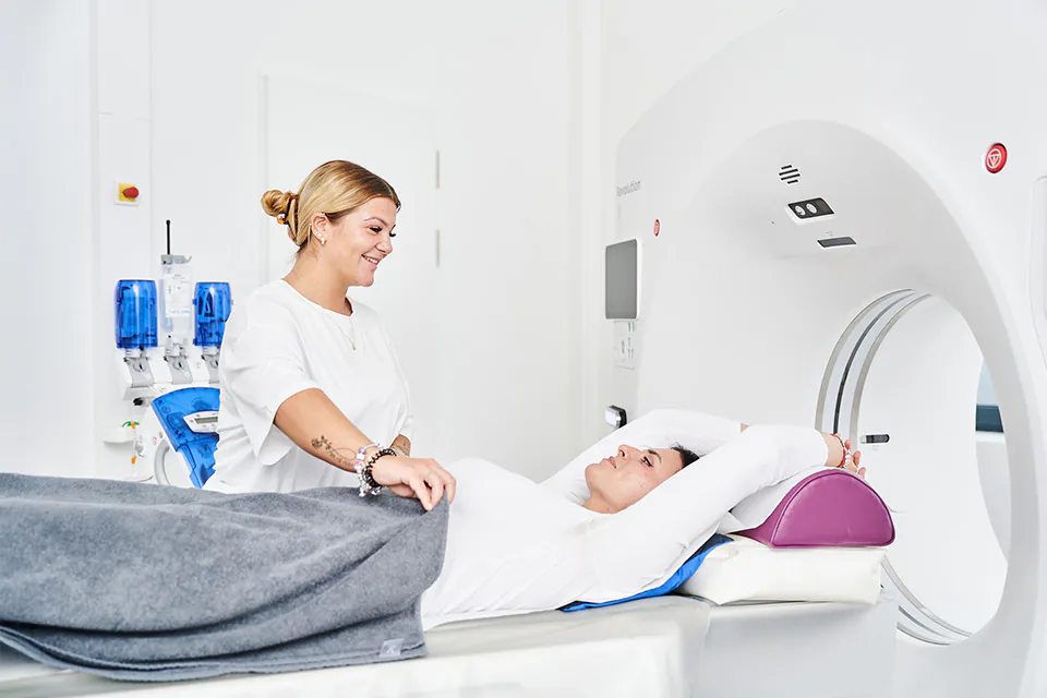 Patientin liegt mit ausgestreckten Armen auf Untersuchungsliege eines CT-Scanners, daneben steht eine medizinische Fachkraft in weißer Kleidung