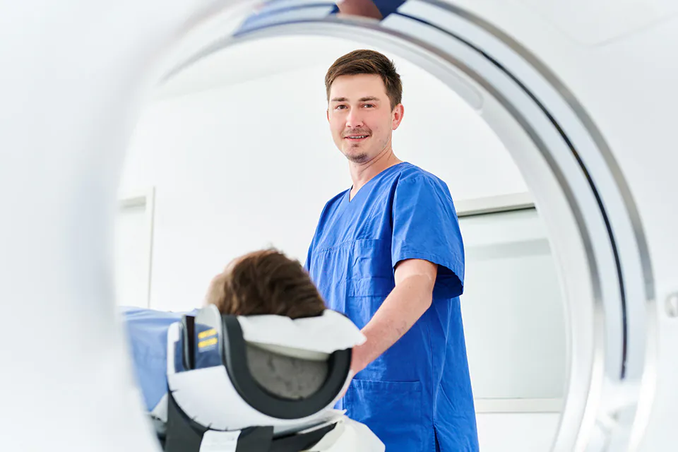 Medizinische Fachkraft in blauer Arbeitskleidung steht neben einer liegenden Person vor einem Gerät für bildgebende Diagnostik.